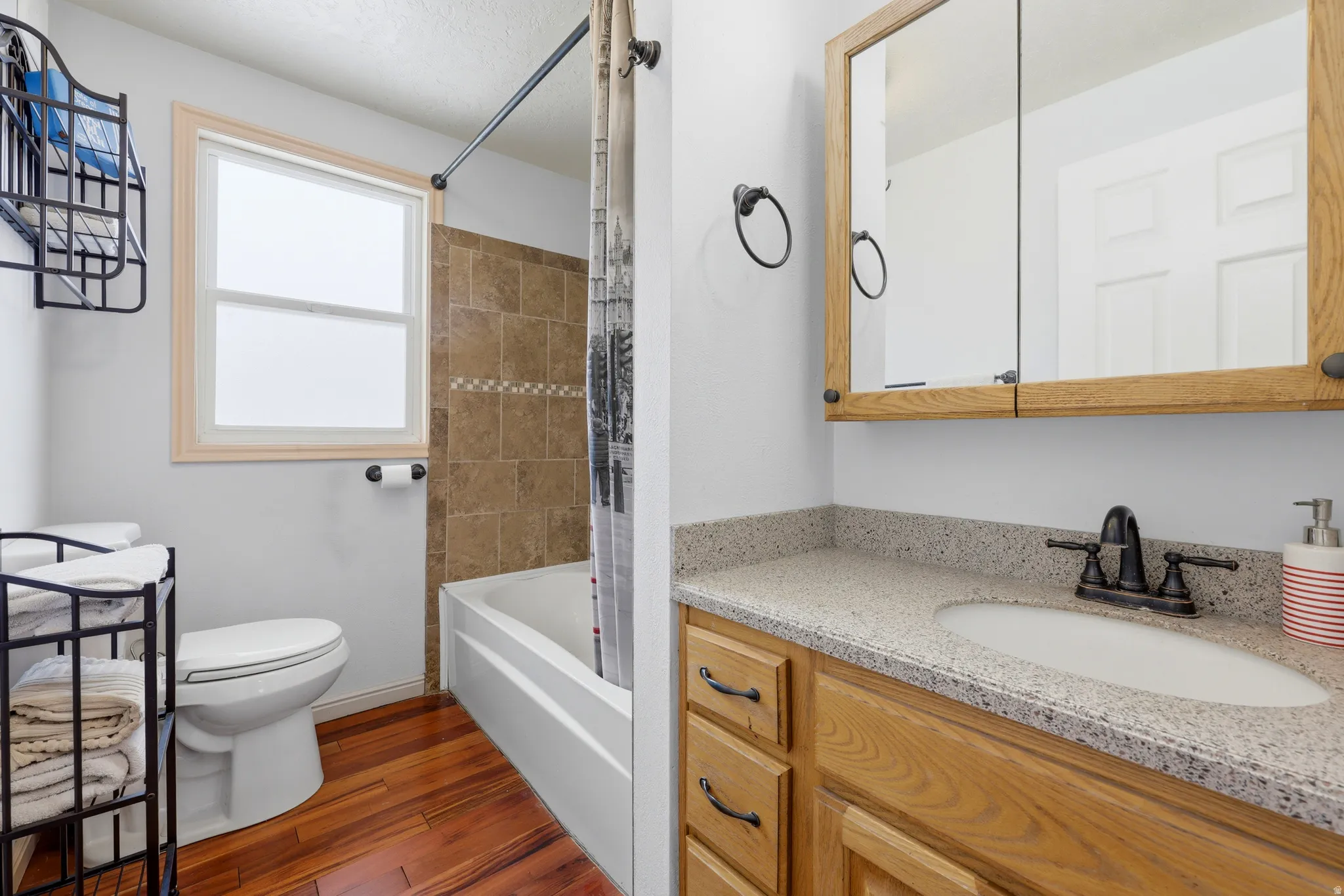 Bathroom featuring wood-finish flooring, a neutral tile tub surround, and a vanity with a light-colored speckled countertop