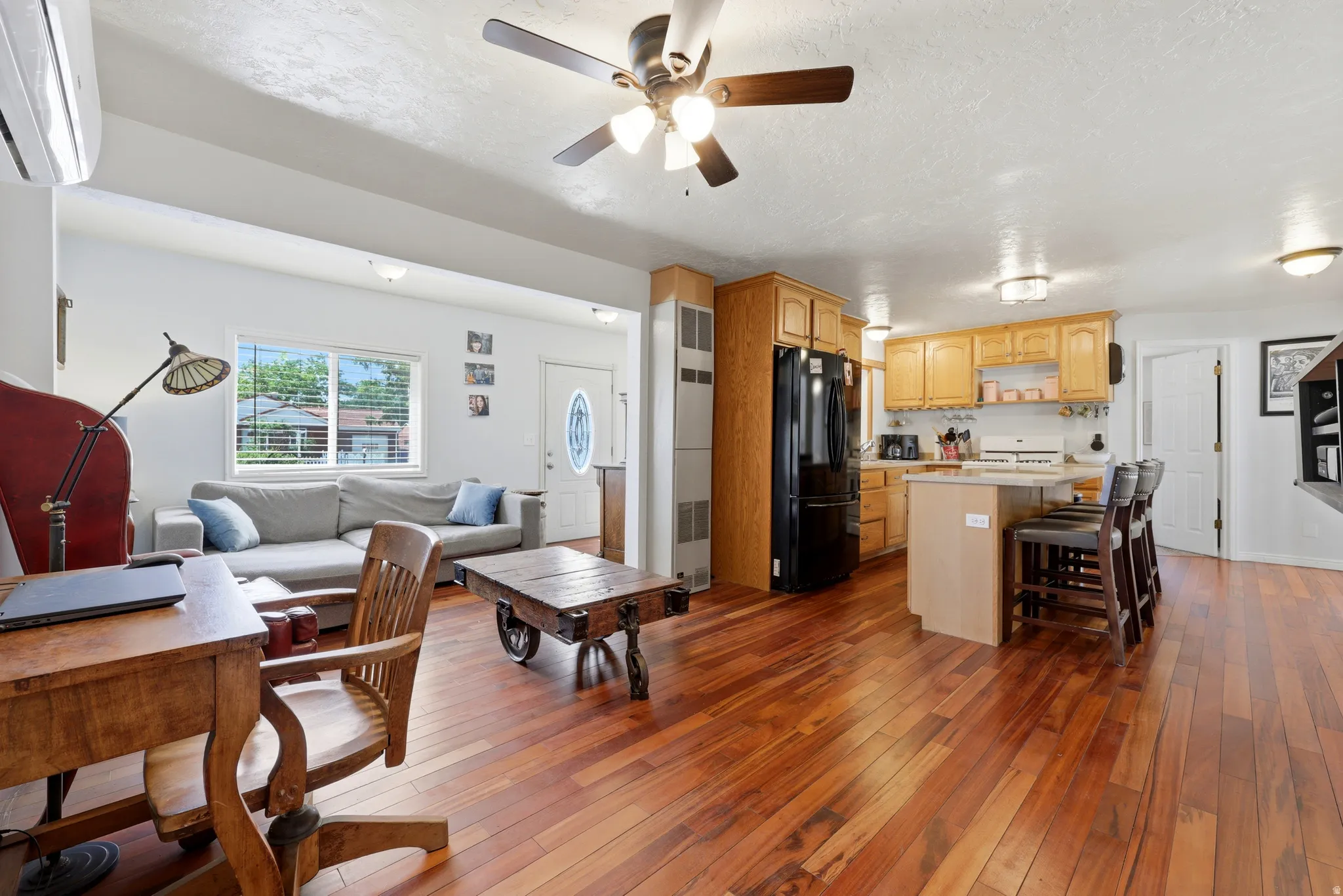 Open-concept living space featuring wood-finish flooring, a kitchen with wood cabinetry and black appliances, a central island with bar seating, and a ceiling fan