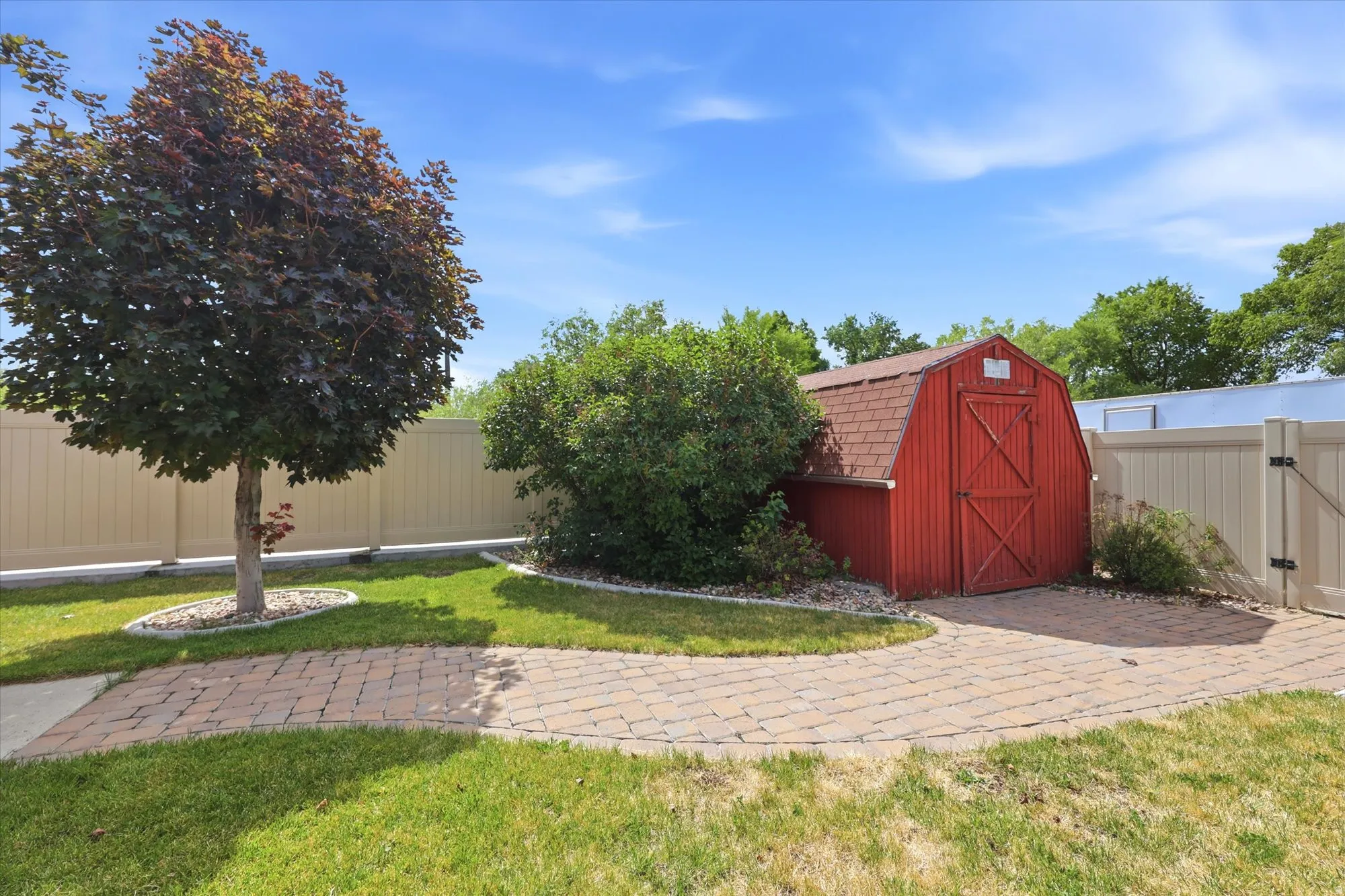 Red barn-style shed with shingle roof, surrounded by established landscaping, a mature tree, and a paver pathway