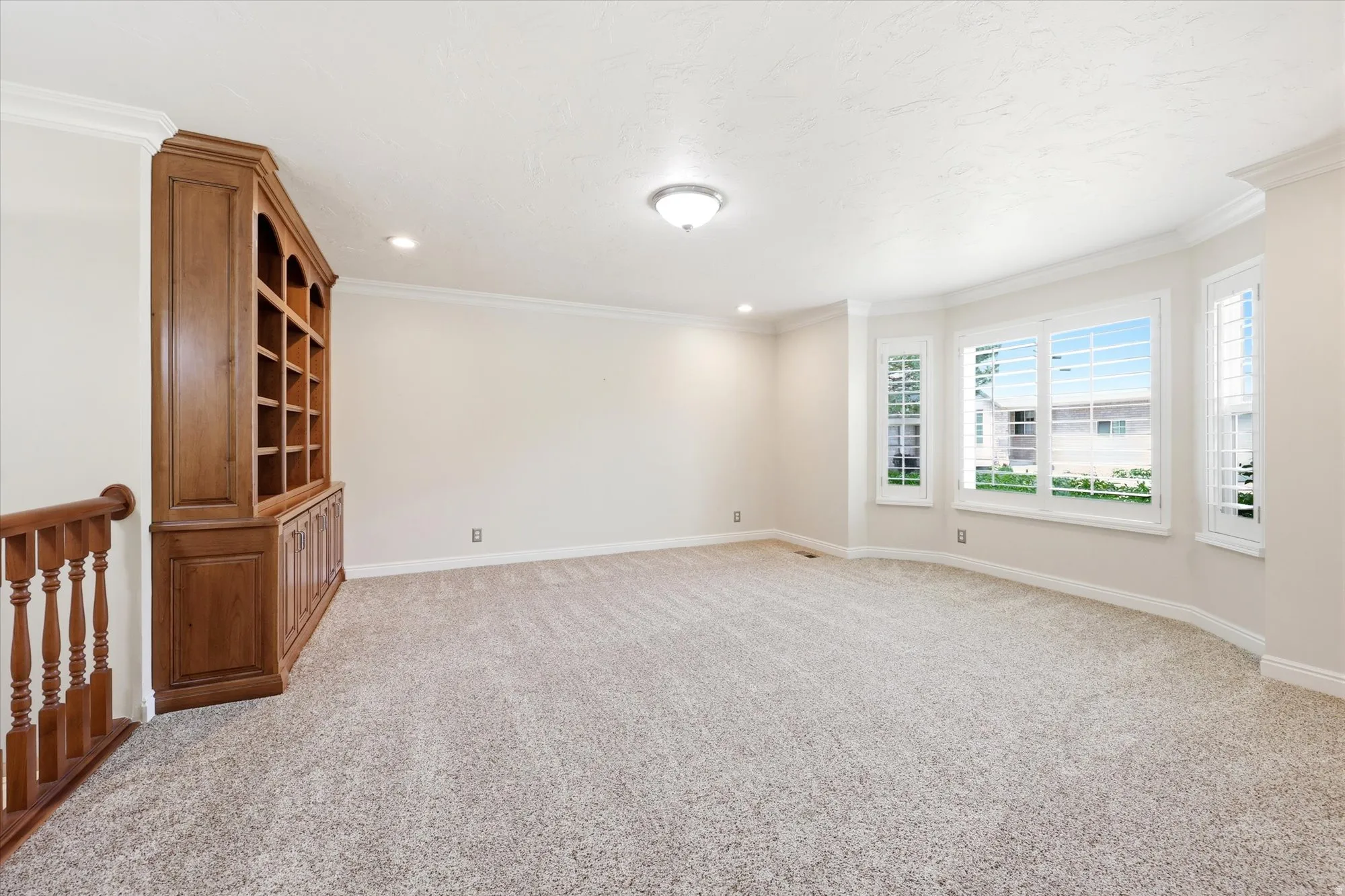 Spacious room featuring a built-in wood-finish bookcase, light-toned carpeting, and a bay window with plantation shutters