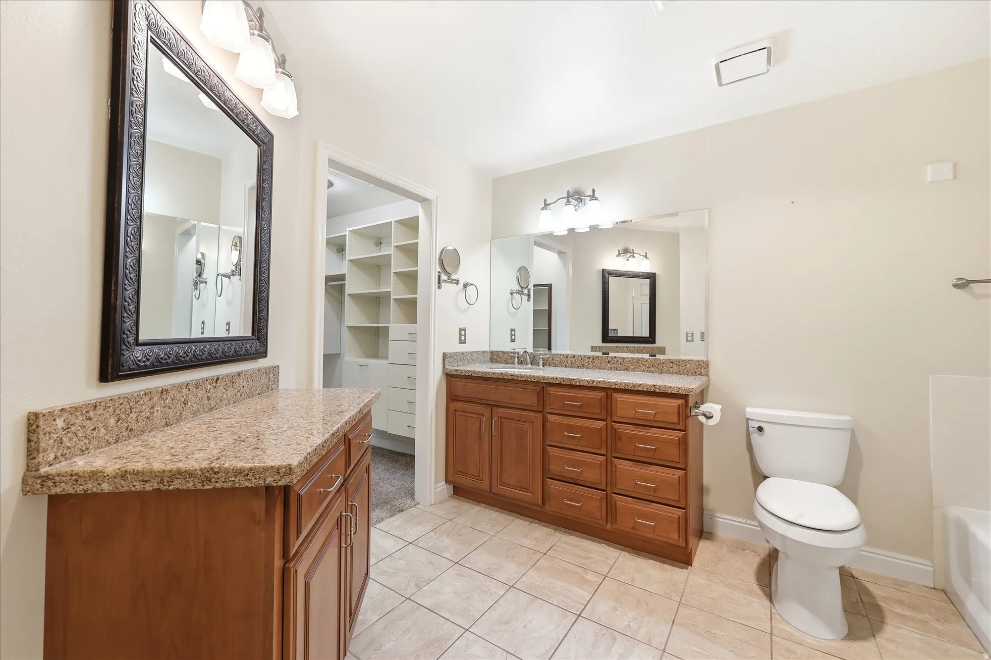 Spacious bathroom featuring dual wood-finish vanities with light-toned countertops, a built-in toilet, and tile flooring