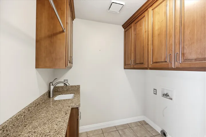 Laundry room featuring extensive wood cabinetry, a utility sink with a stone countertop, a built-in hanging rod, and tile flooring