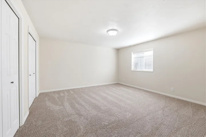 Spacious room featuring neutral carpeting, light-colored walls, a ceiling-mounted light fixture, a window with horizontal blinds, and two panel doors with black hardware