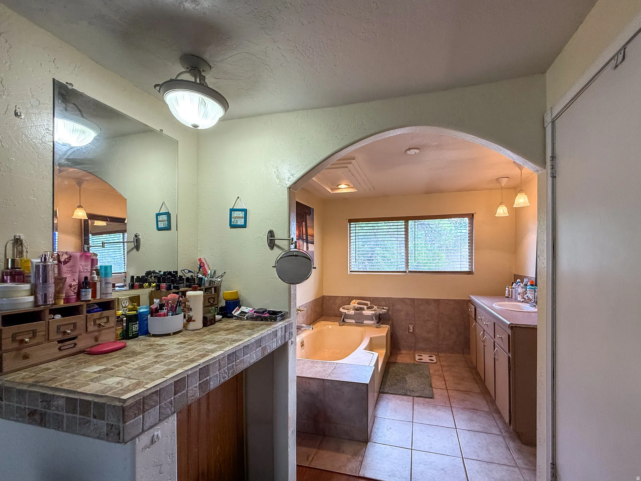 Bathroom featuring a tiled vanity countertop, large wall mirror, arched doorway, built-in bathtub, and dual windows with blinds