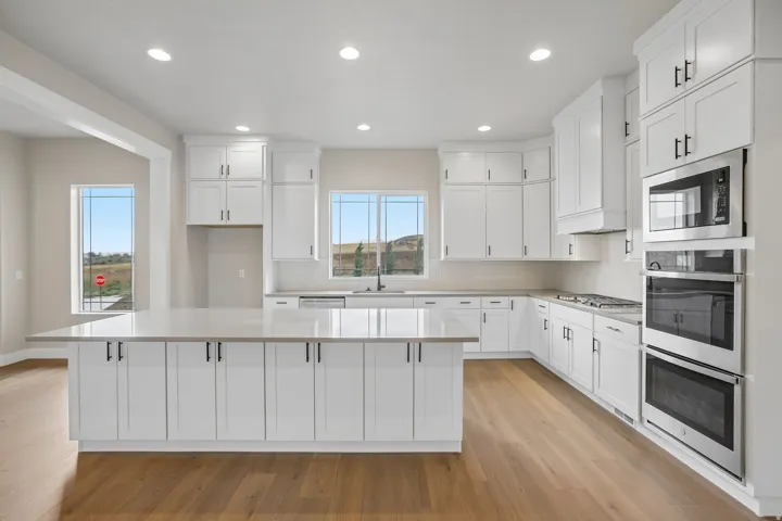 Contemporary kitchen featuring white shaker cabinetry, an expansive central island with stone-finish countertop, wood-finish flooring, stainless steel built-in appliances, and recessed lighting