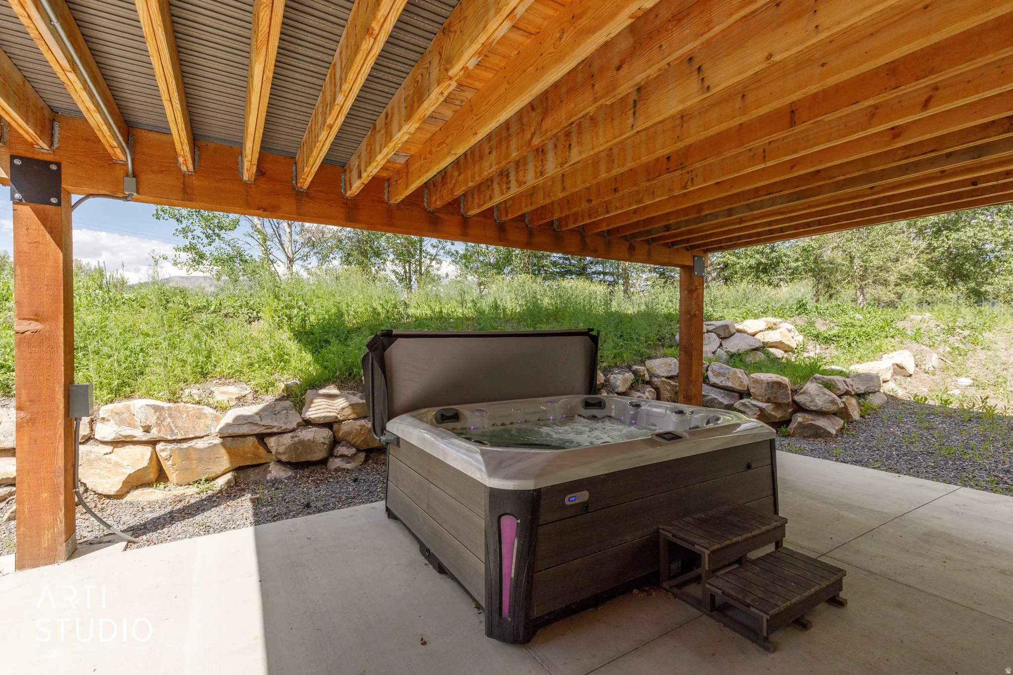 Outdoor spa area featuring a bubbling hot tub with wooden steps, set on a concrete patio under a wooden deck with a corrugated metal ceiling