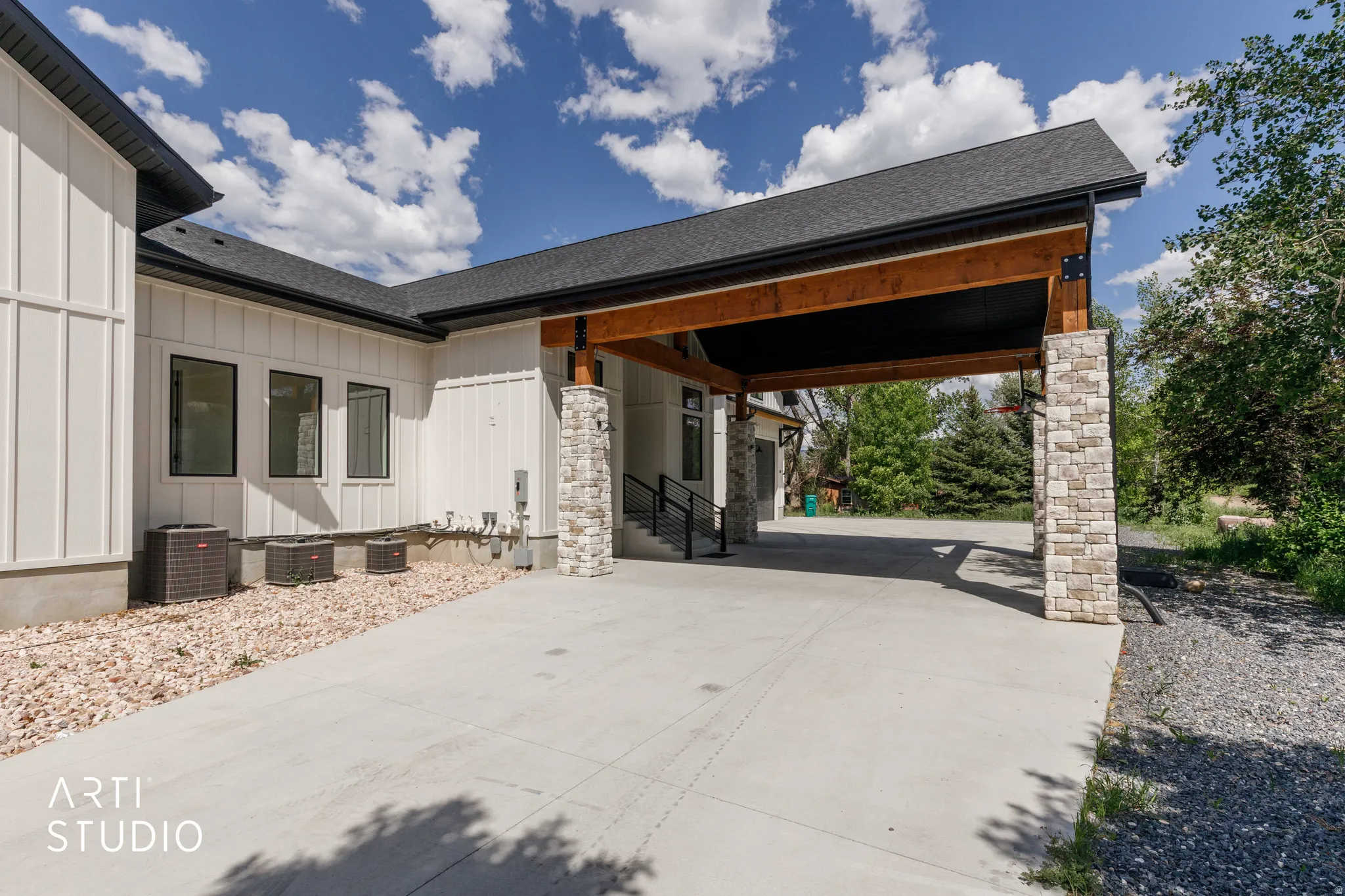 Covered carport featuring exposed wood beams and stacked stone columns, complemented by a board and batten exterior, dark-framed windows, and a gravel landscape bed