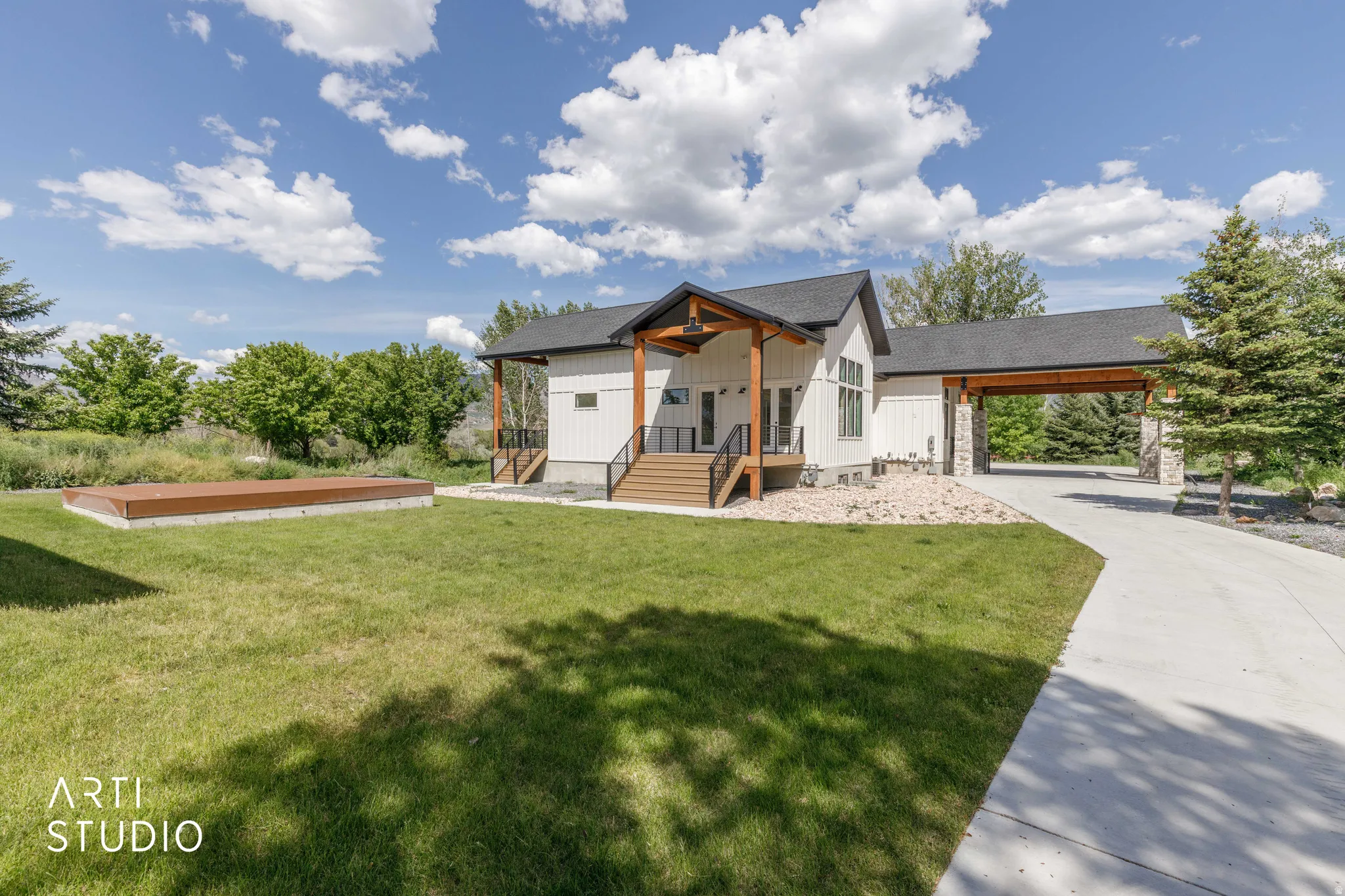 Modern farmhouse exterior featuring white vertical siding, a dark pitched roof, wood-finish columns, a covered front porch with wood-finish steps, and a concrete driveway leading to a covered carport with stone pillar supports