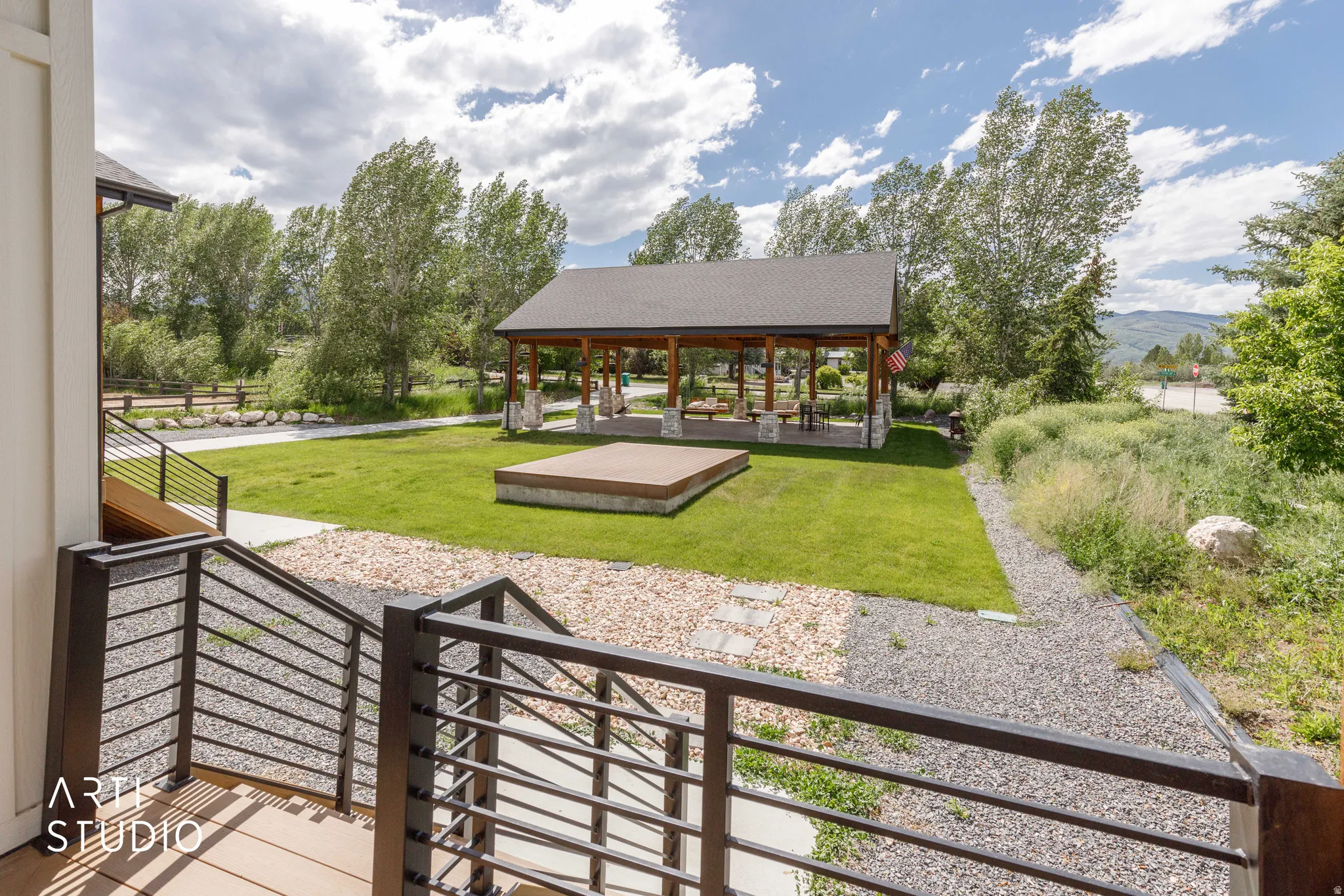 Outdoor gathering area featuring a covered pavilion with stone pillars, a wooden stage, and a manicured lawn