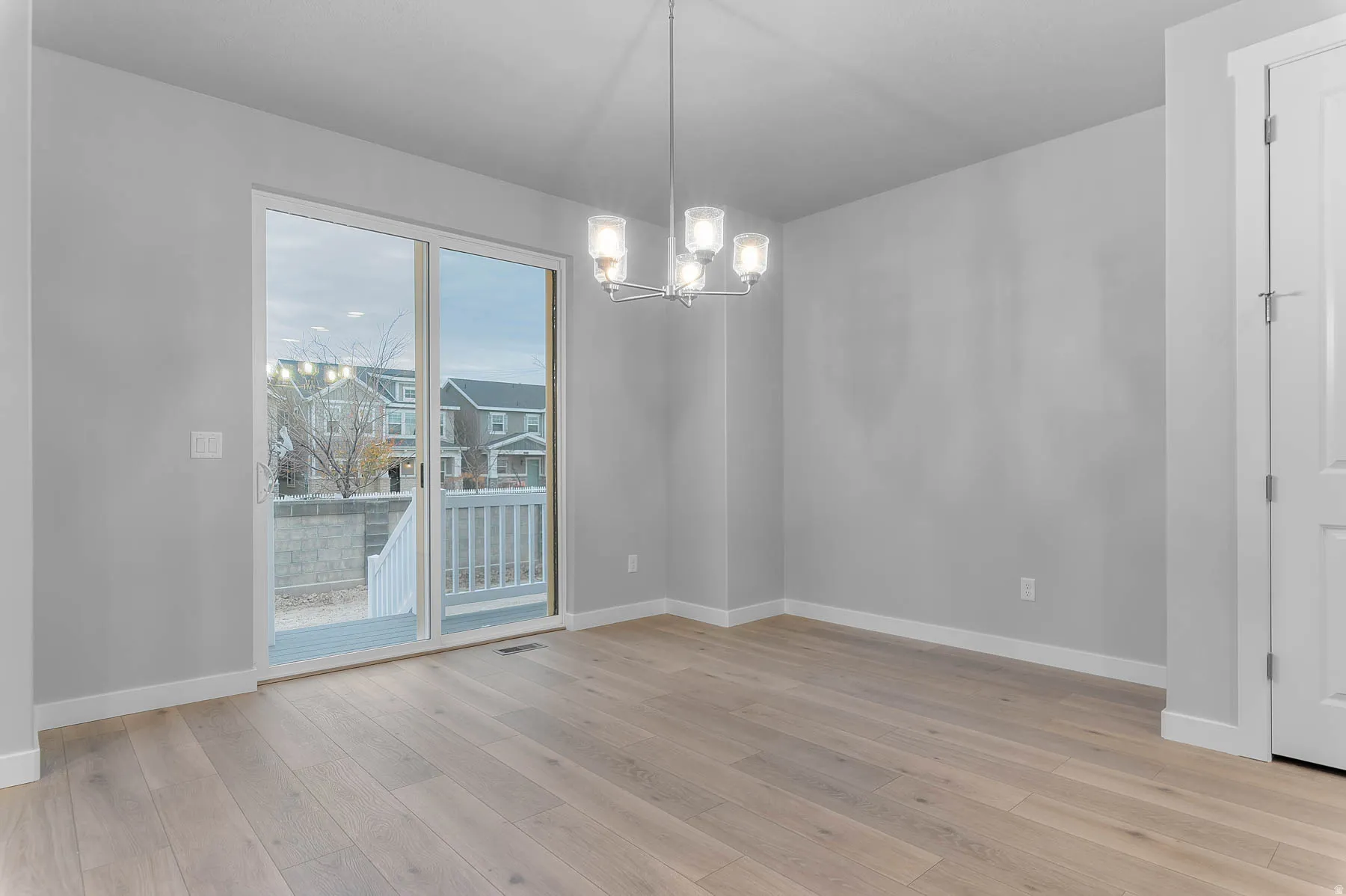 Spacious room featuring light wood-finish flooring, a contemporary chandelier, white baseboards, and a glass sliding door leading to an outdoor deck with white railings