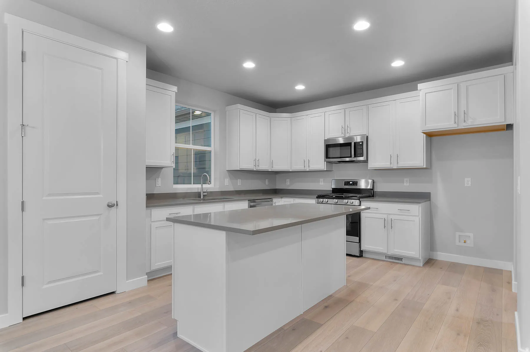 Contemporary kitchen featuring light wood-finish flooring, white cabinetry, a central island with a grey countertop, stainless steel appliances, and recessed lighting