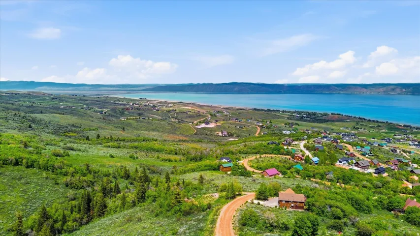 Expansive hillside landscape featuring a tranquil body of water, rolling green terrain, scattered residential structures, and a winding unpaved road