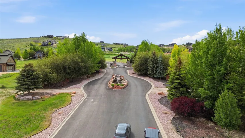 Paved entry drive with a central stone-clad water feature, bordered by mature trees and landscaped gravel beds