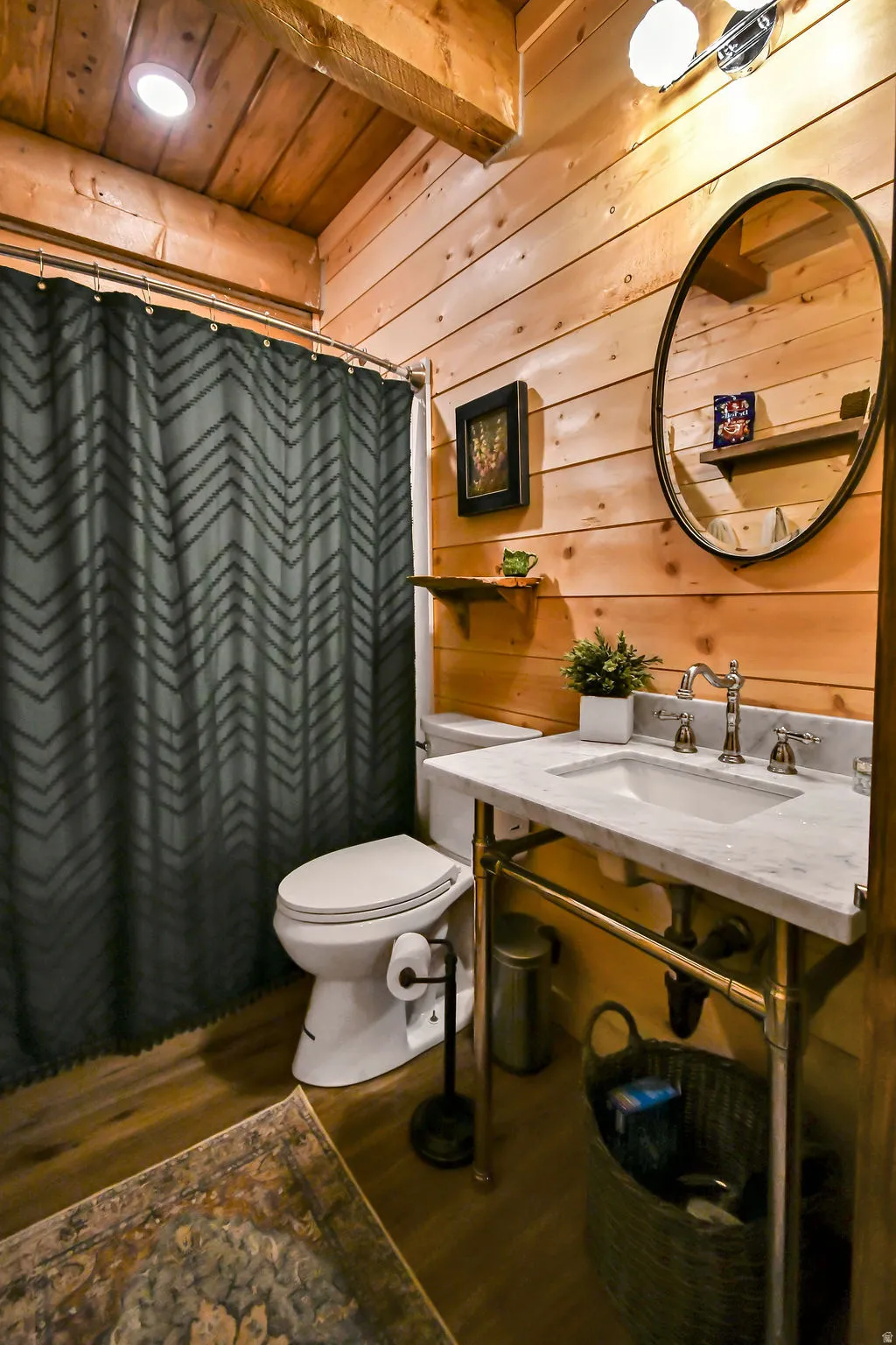 Bathroom featuring wood plank walls, exposed wood ceiling beams, wood-finish flooring, a white vanity with a marble-look countertop, and a circular mirror