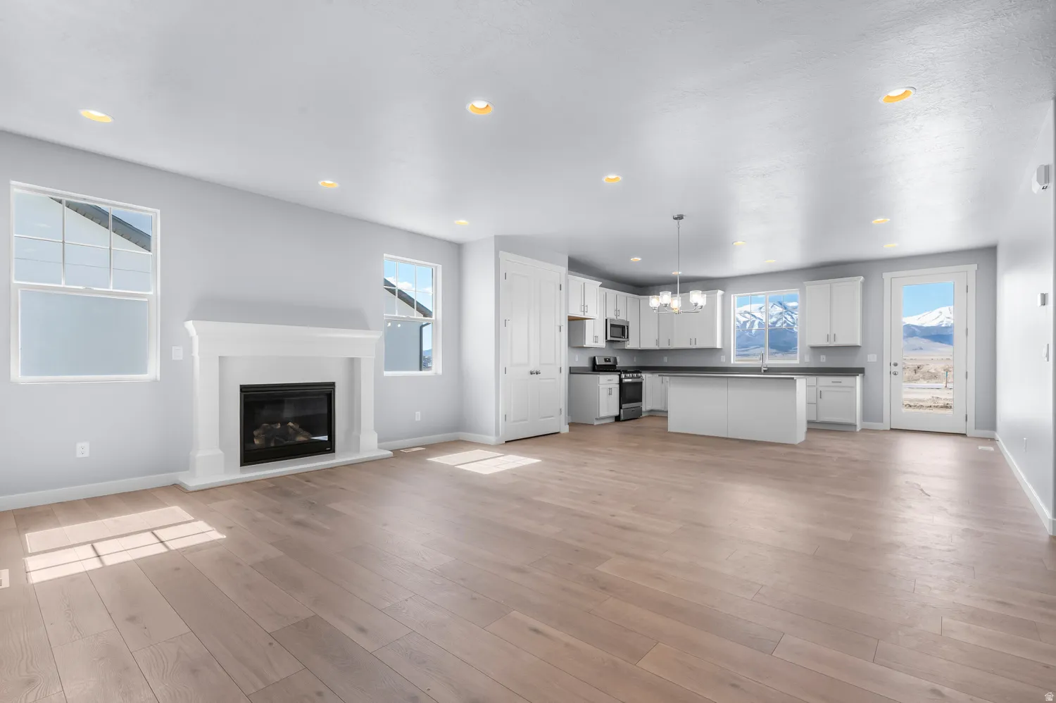Open-concept living area featuring light wood-finish flooring, a white mantel fireplace, recessed lighting, and a kitchen with white cabinetry and dark countertops