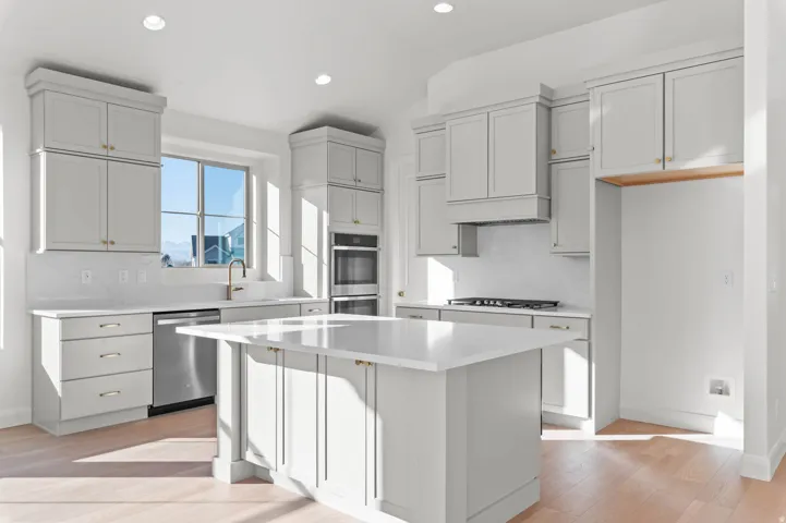 Modern kitchen featuring light wood-finish flooring, white cabinetry with gold-tone hardware, a central island with a white countertop, and stainless steel appliances including a dishwasher, gas range, and double wall ovens