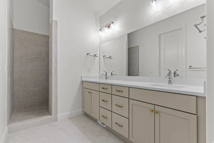 Spacious bathroom featuring a dual vanity with light-toned cabinetry and gold-toned hardware, solid surface countertops, a full-width mirror, and an adjacent walk-in shower with tiled walls and flooring