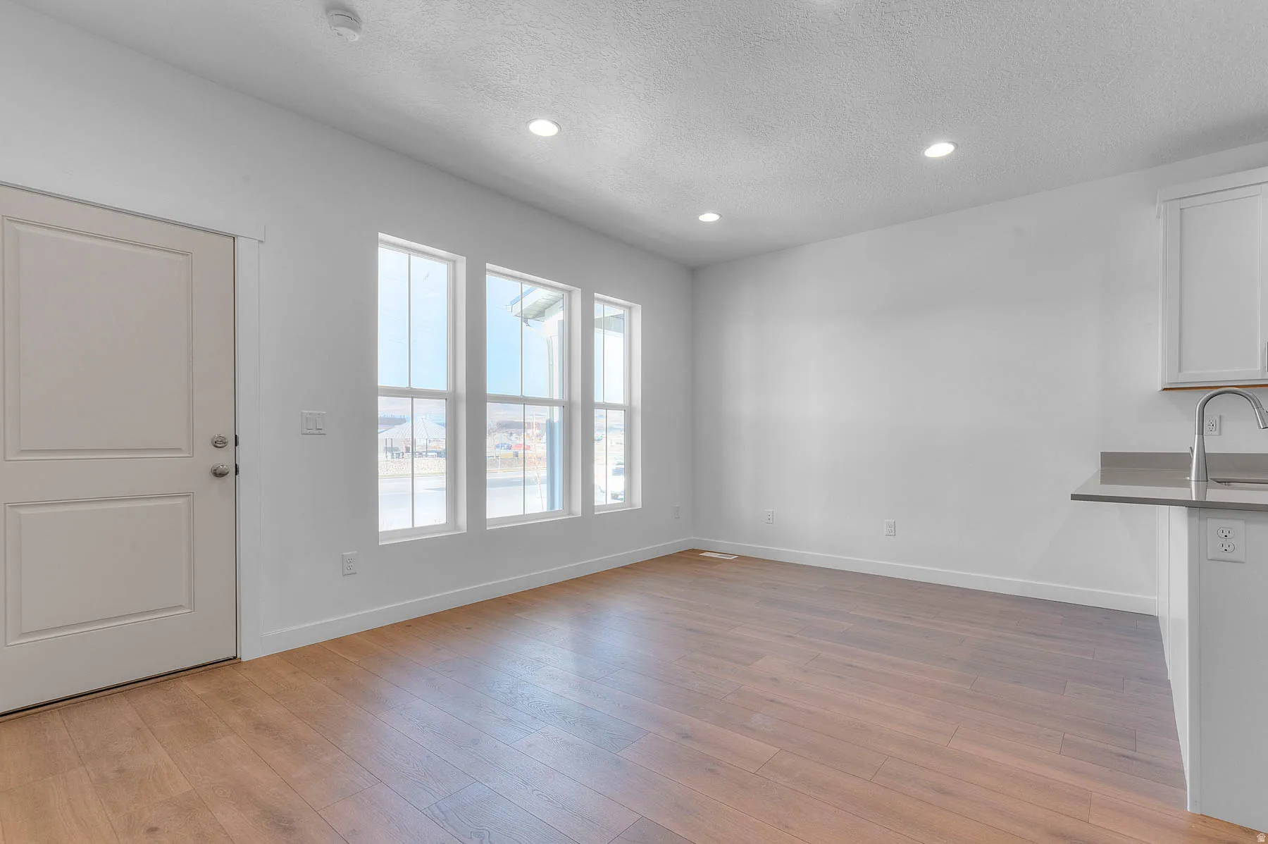 Open concept living space featuring wood-finish flooring, a white paneled door, three vertical windows, recessed lighting, and a kitchen peninsula with a stainless steel faucet and gray countertop
