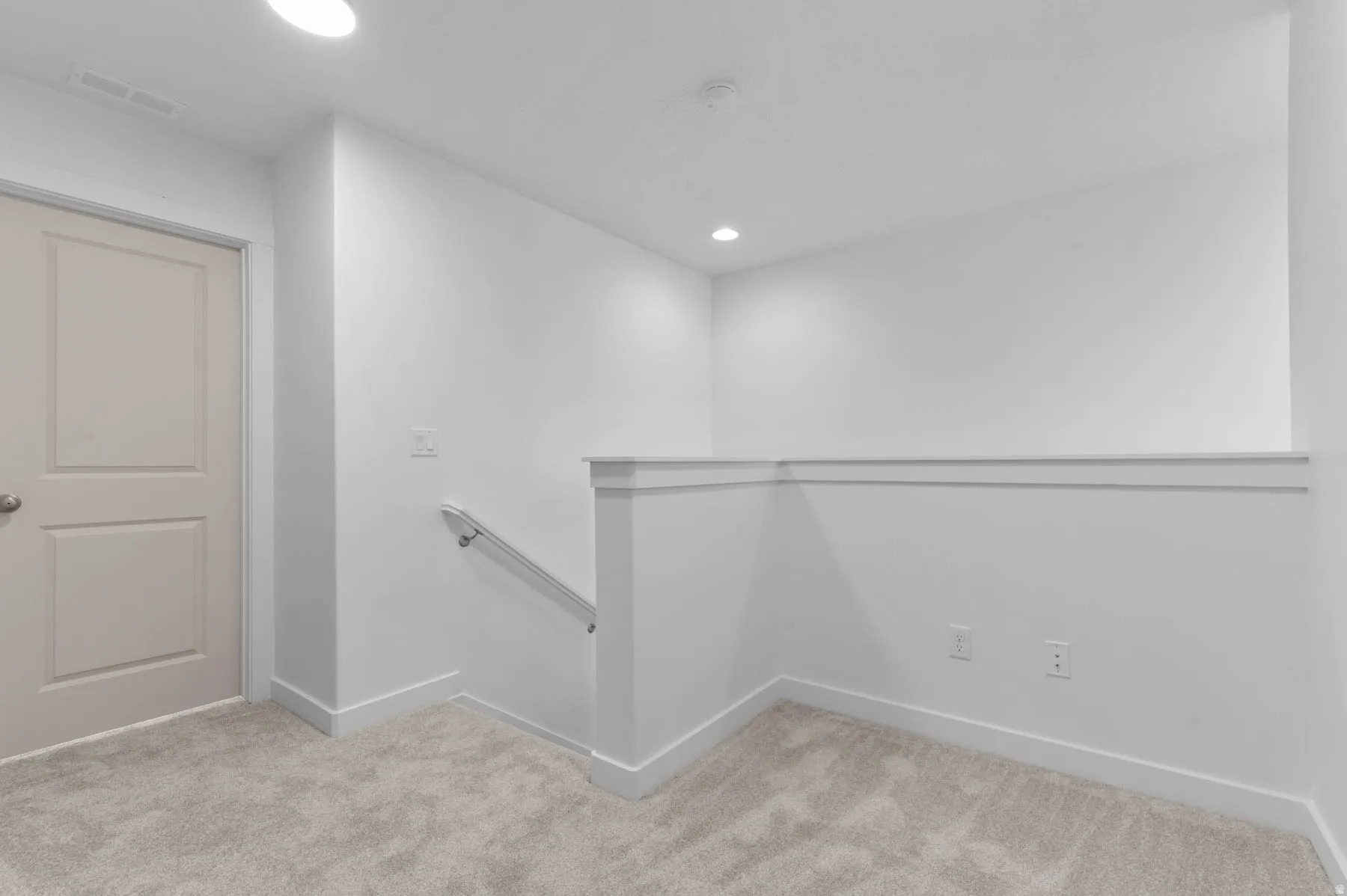 Carpeted landing area featuring a neutral panel door, white walls, recessed lighting, and a white handrail on a half-wall partition