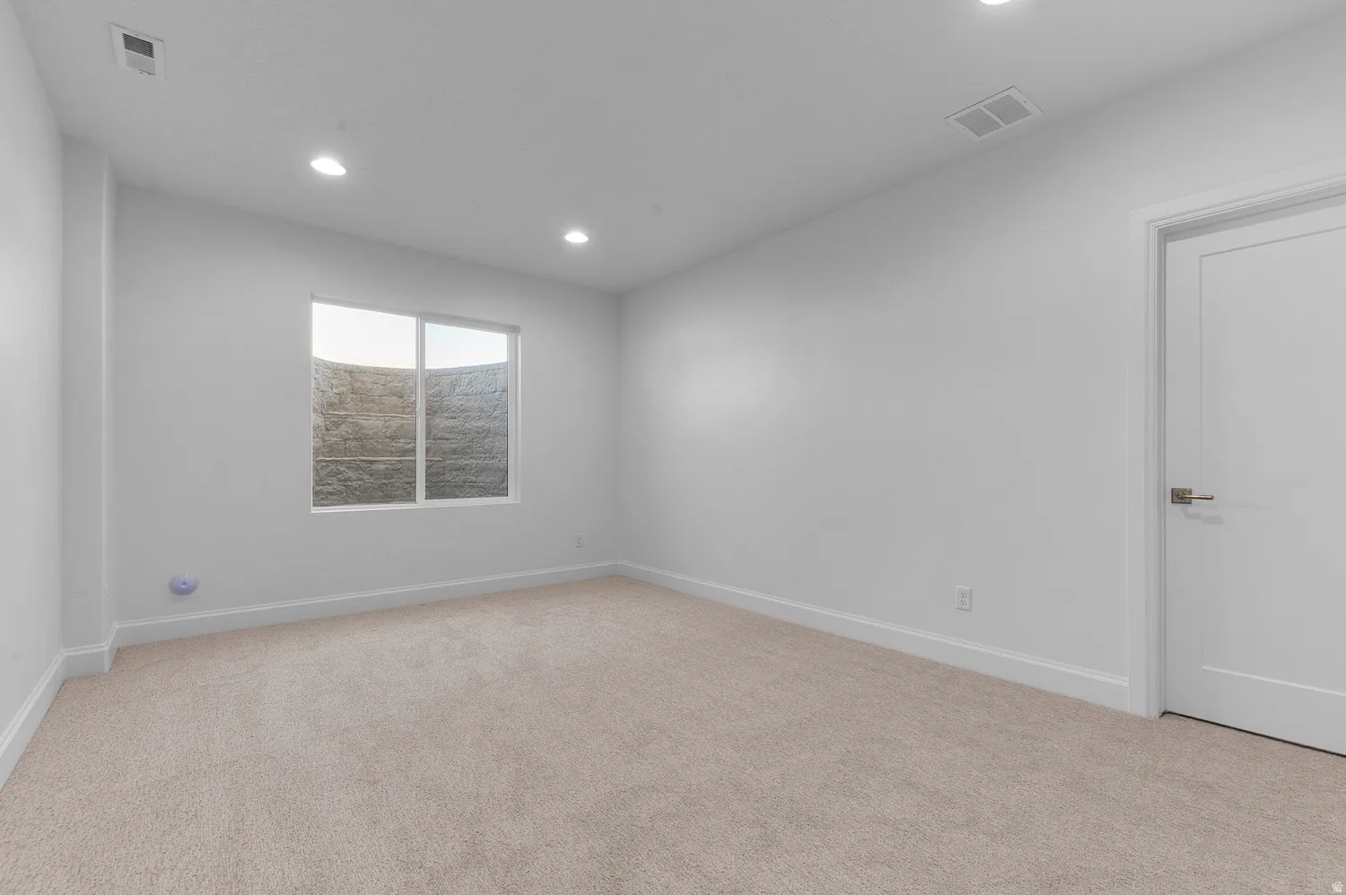 Bright room featuring soft carpet flooring, recessed ceiling lighting, a white painted door with contemporary hardware, and a window with a stone-textured exterior wall