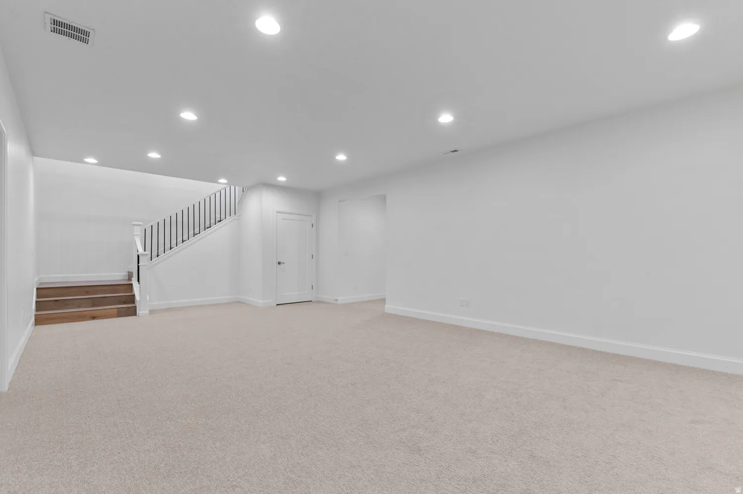 Spacious lower-level room featuring light-toned carpeting, recessed ceiling lighting, a staircase with wood-finish treads and black balusters, and a white interior door