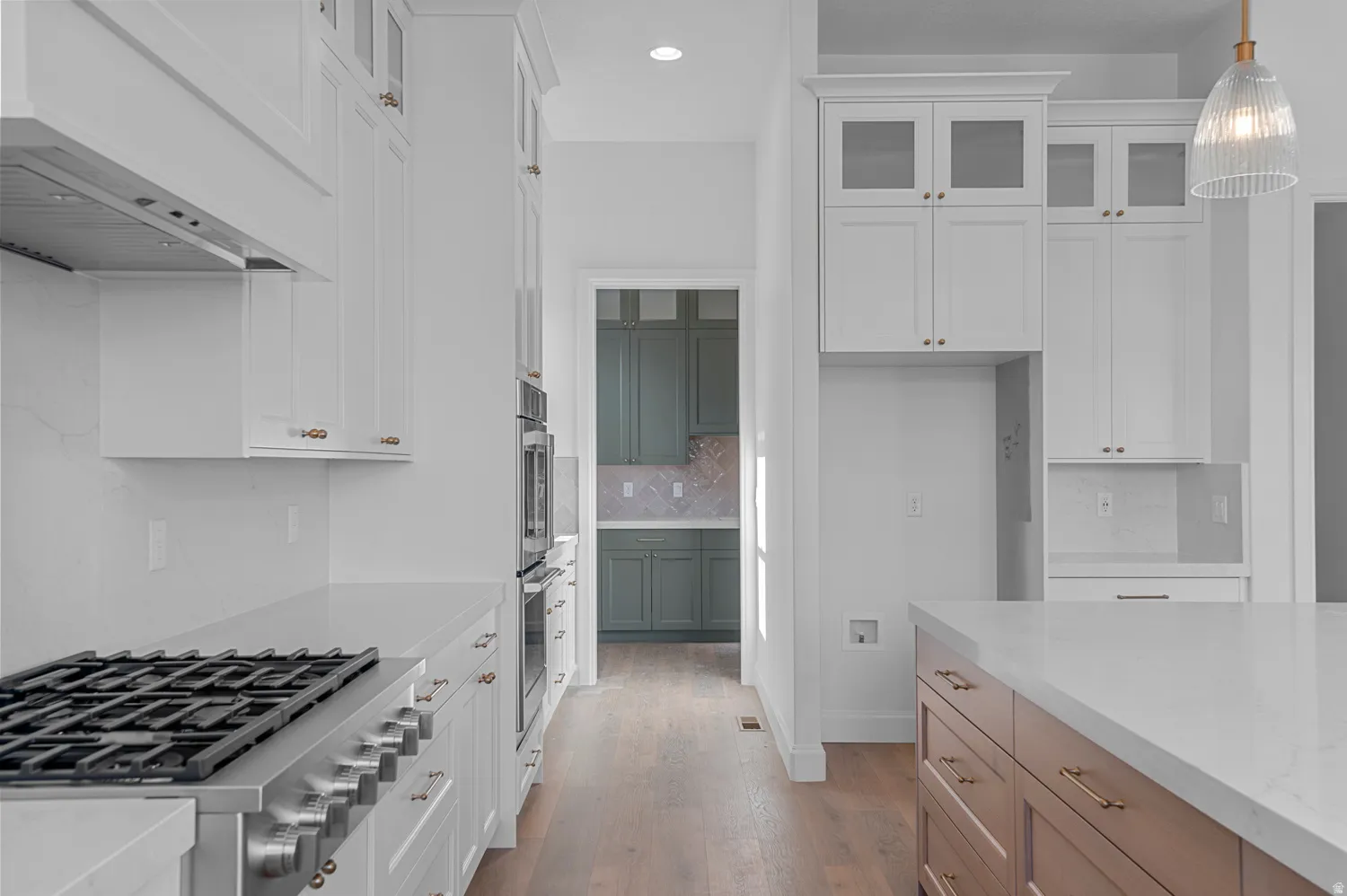 Gourmet kitchen featuring a stainless steel gas range, built-in double ovens, white shaker cabinetry, an expansive island with a light-colored countertop, and wood-finish flooring