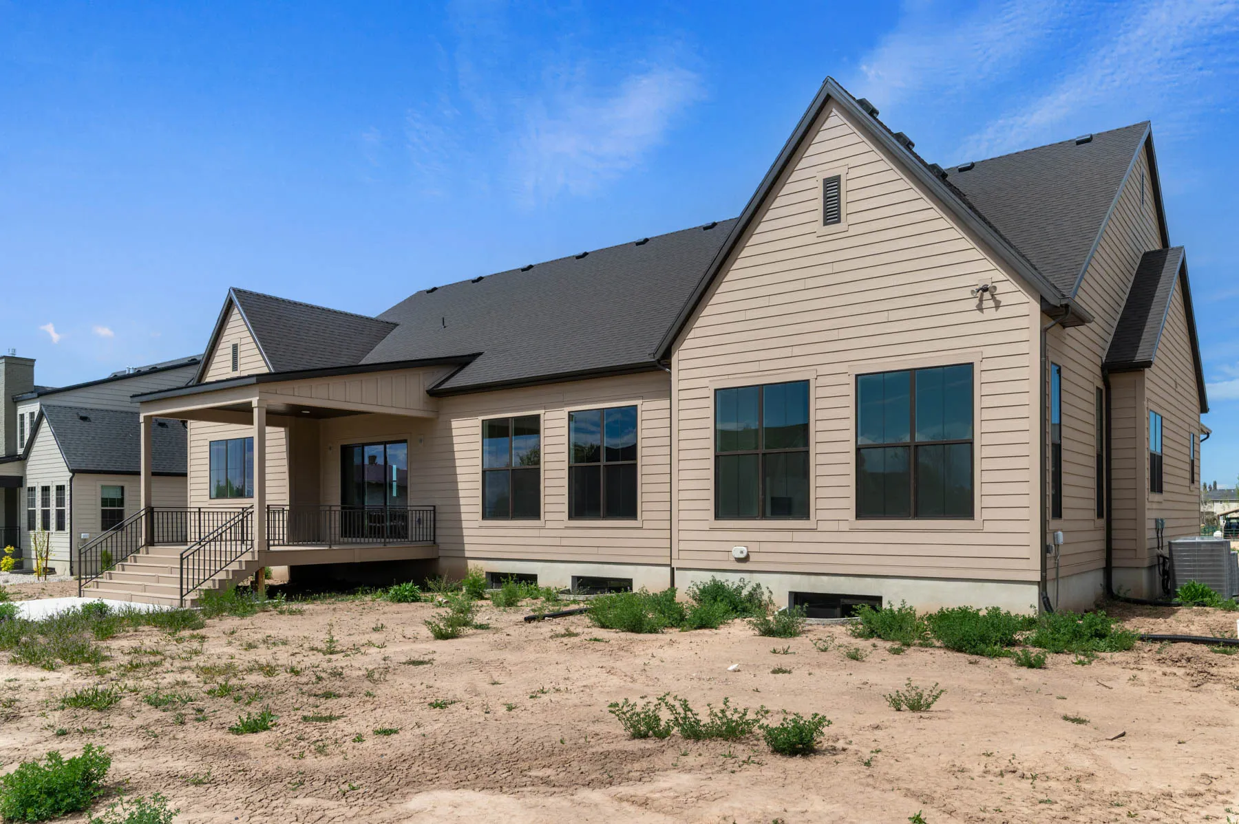 Rear exterior featuring a covered patio with dark railing, multiple large windows, tan horizontal siding, and a dark shingle roof