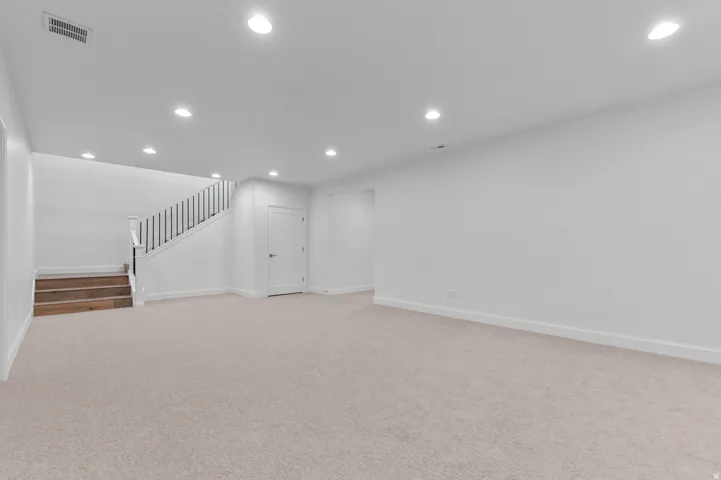 Spacious lower-level room featuring light-toned carpeting, recessed ceiling lighting, a staircase with wood-finish treads and black balusters, and a white interior door