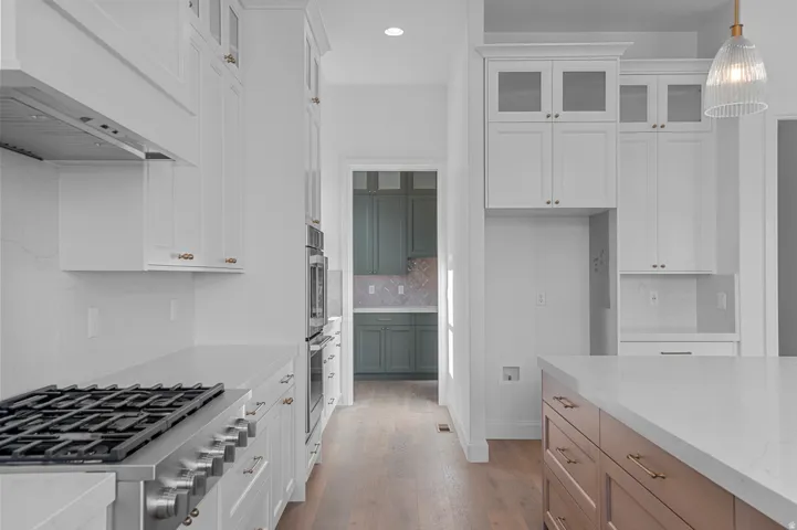 Gourmet kitchen featuring a stainless steel gas range, built-in double ovens, white shaker cabinetry, an expansive island with a light-colored countertop, and wood-finish flooring