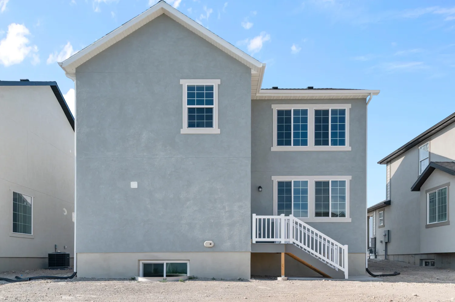 Rear exterior featuring light grey stucco siding, white window trim, multiple large windows, a white-railed staircase, and a visible basement window