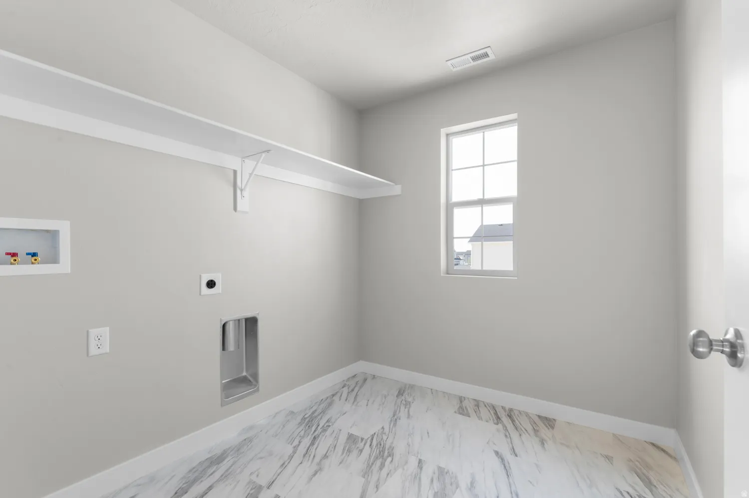 Utility room featuring a single window, a long white storage shelf, pre-installed washer and dryer hookups, and large format marble-look tile flooring
