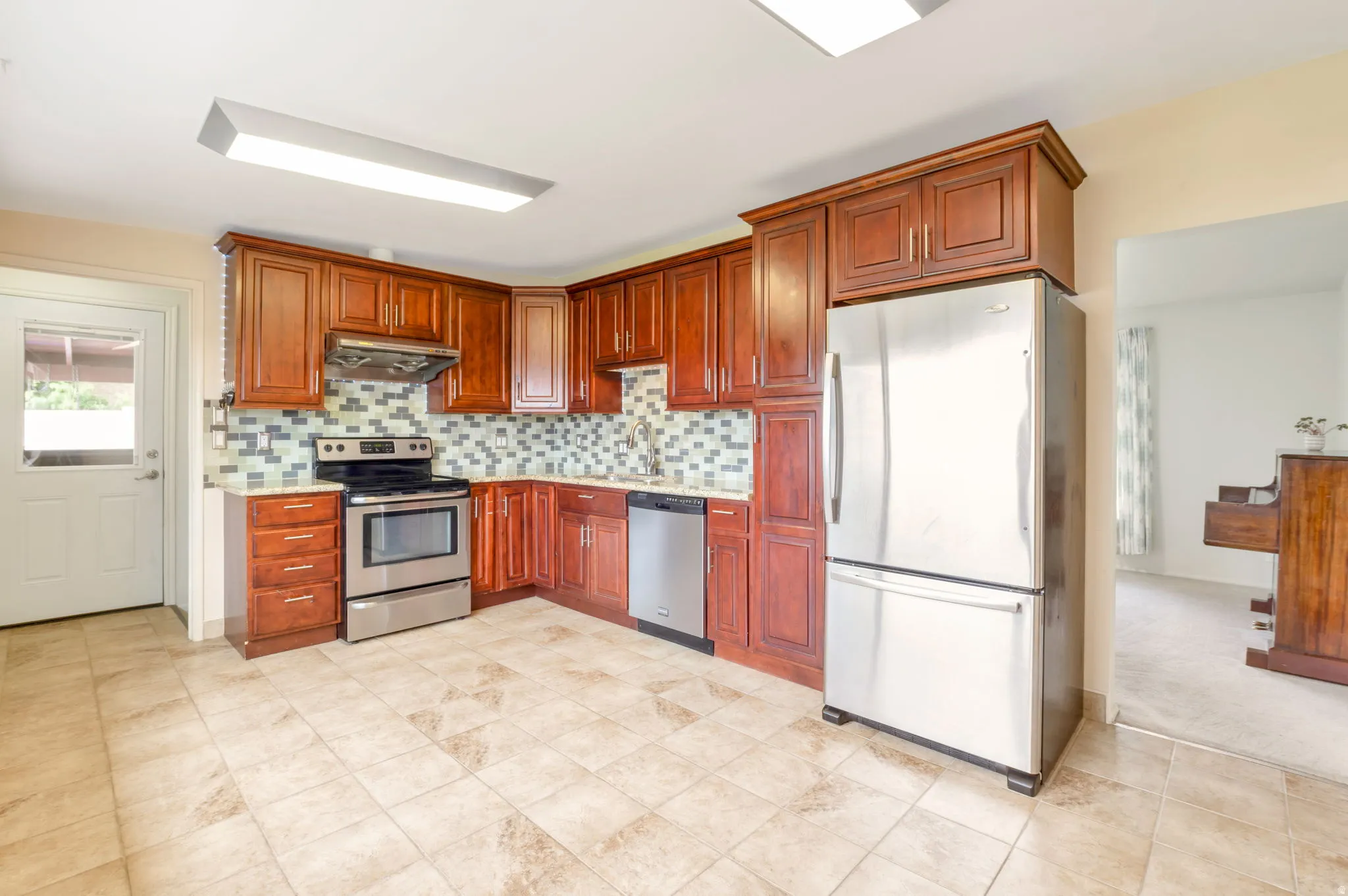 Kitchen featuring rich wood cabinetry, stainless steel appliances, a mosaic tile backsplash, and light tile flooring