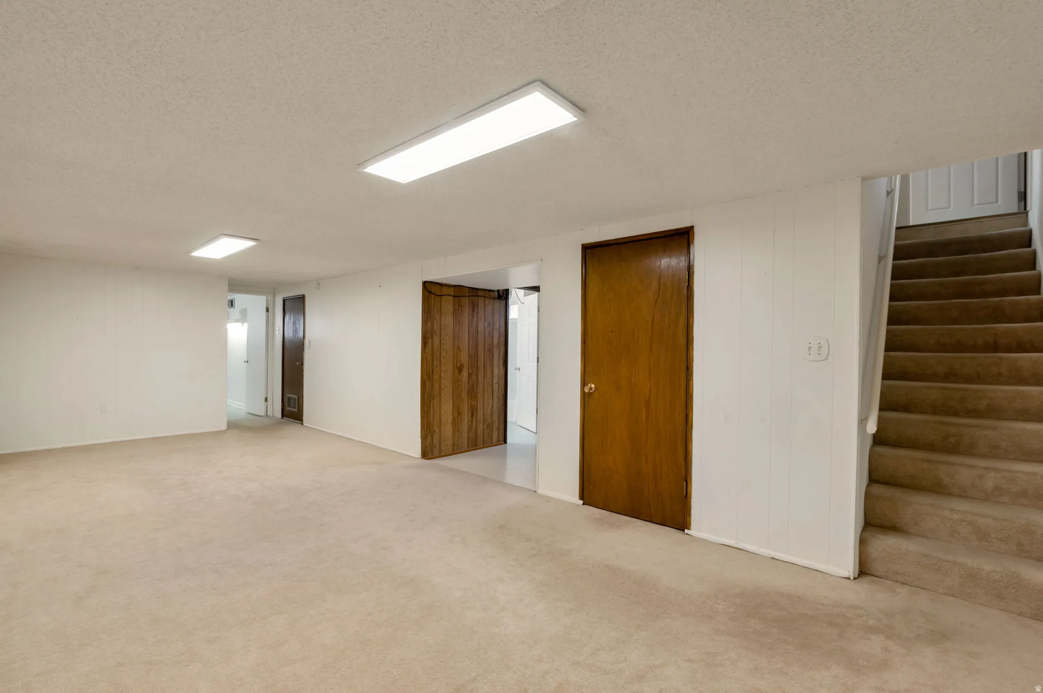 Spacious carpeted room featuring white walls, flush-mount ceiling lighting, and a staircase with matching carpet