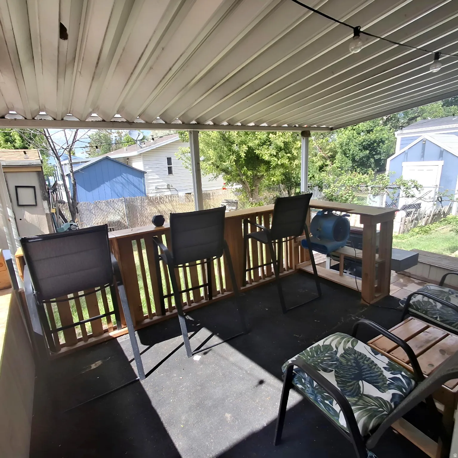 Covered outdoor patio featuring a slatted roof, integrated wooden bar with railing, string lighting, and a dark-toned floor surface