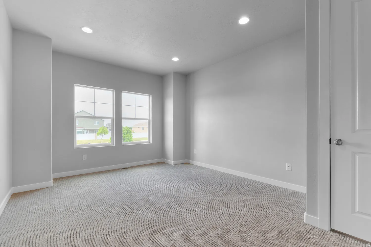 Neutral tone wall paint, recessed ceiling lighting, light gray patterned carpet, white baseboards, and two large window panes