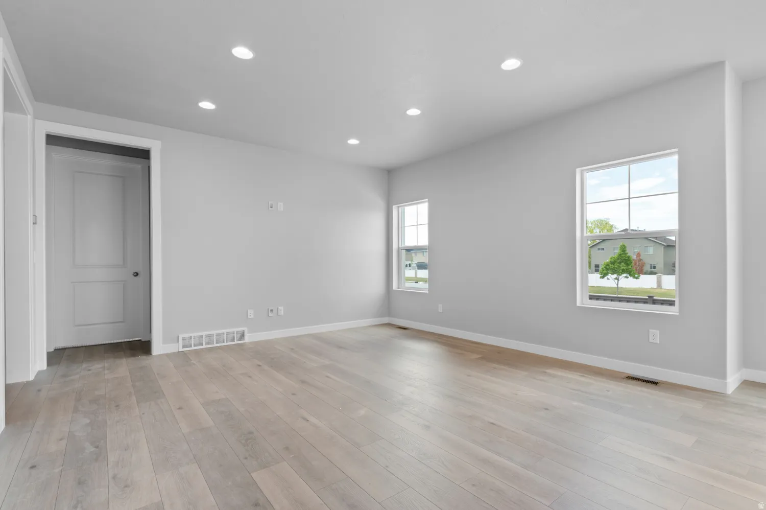 Spacious room featuring light wood-finish flooring, recessed ceiling lighting, two large windows, and a panel door