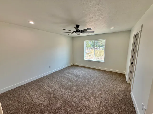 Spacious bedroom featuring neutral wall finishes, recessed ceiling lighting, a ceiling fan with dark blades, carpeting in warm earth tones, and a large window with natural light exposure and exterior views