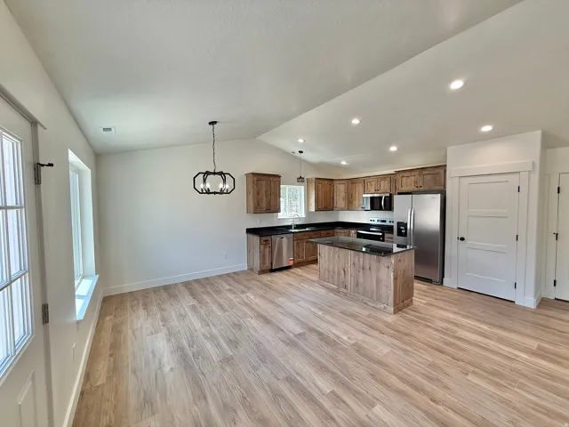 Open-concept kitchen and dining area featuring wood-finish flooring throughout, stainless steel appliances, black countertops, wood cabinetry with an island, recessed lighting, and a pendant fixture