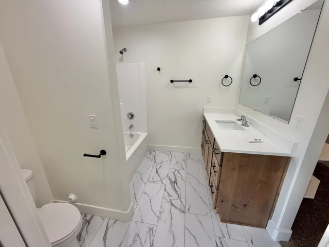 White walls, wood-finish vanity with a white countertop, large wall-mounted mirror, and a white tiled shower/tub combination