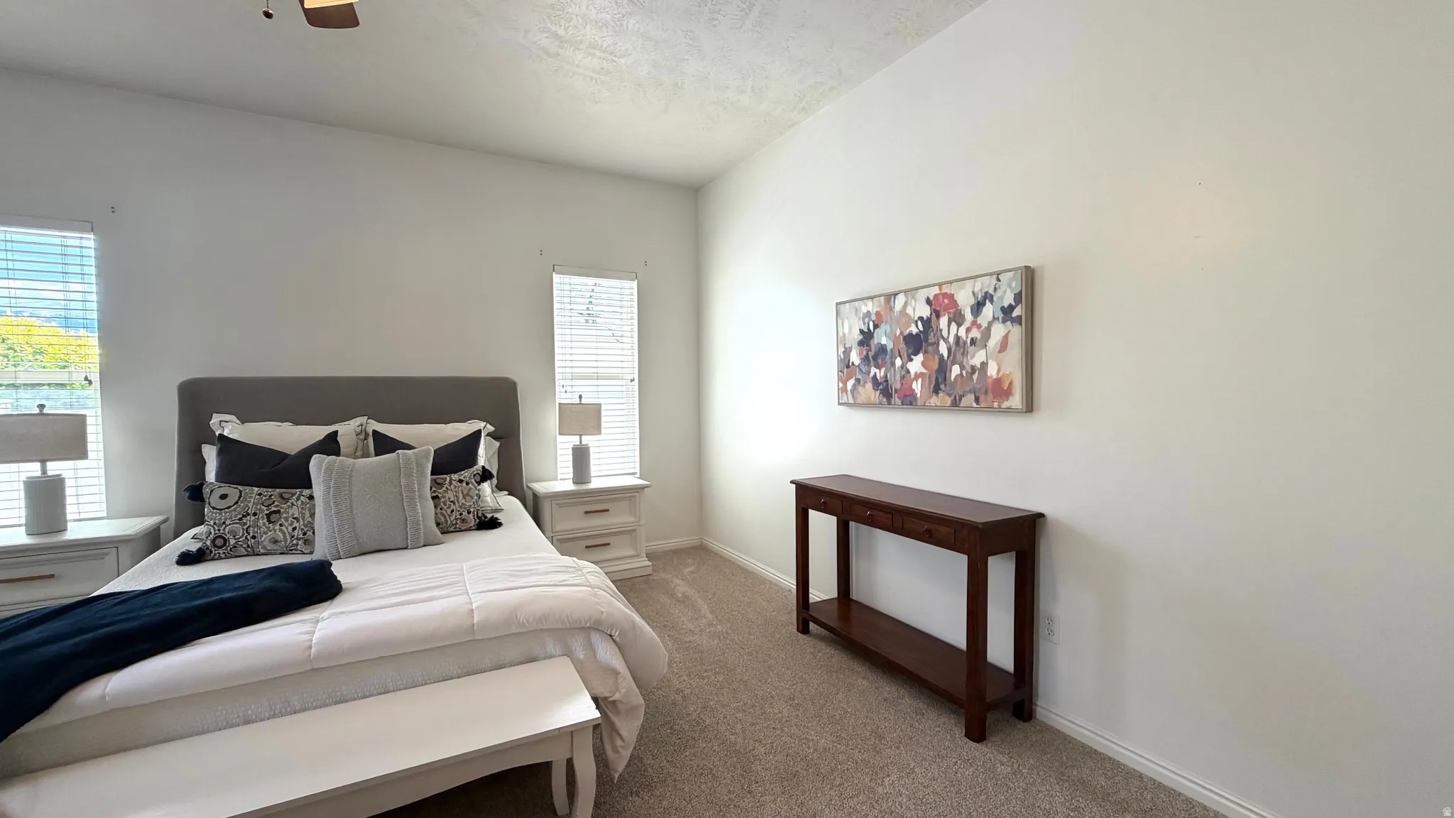 Carpeted room featuring a ceiling fan, dual windows with blinds, a prominent upholstered headboard, and light-colored walls