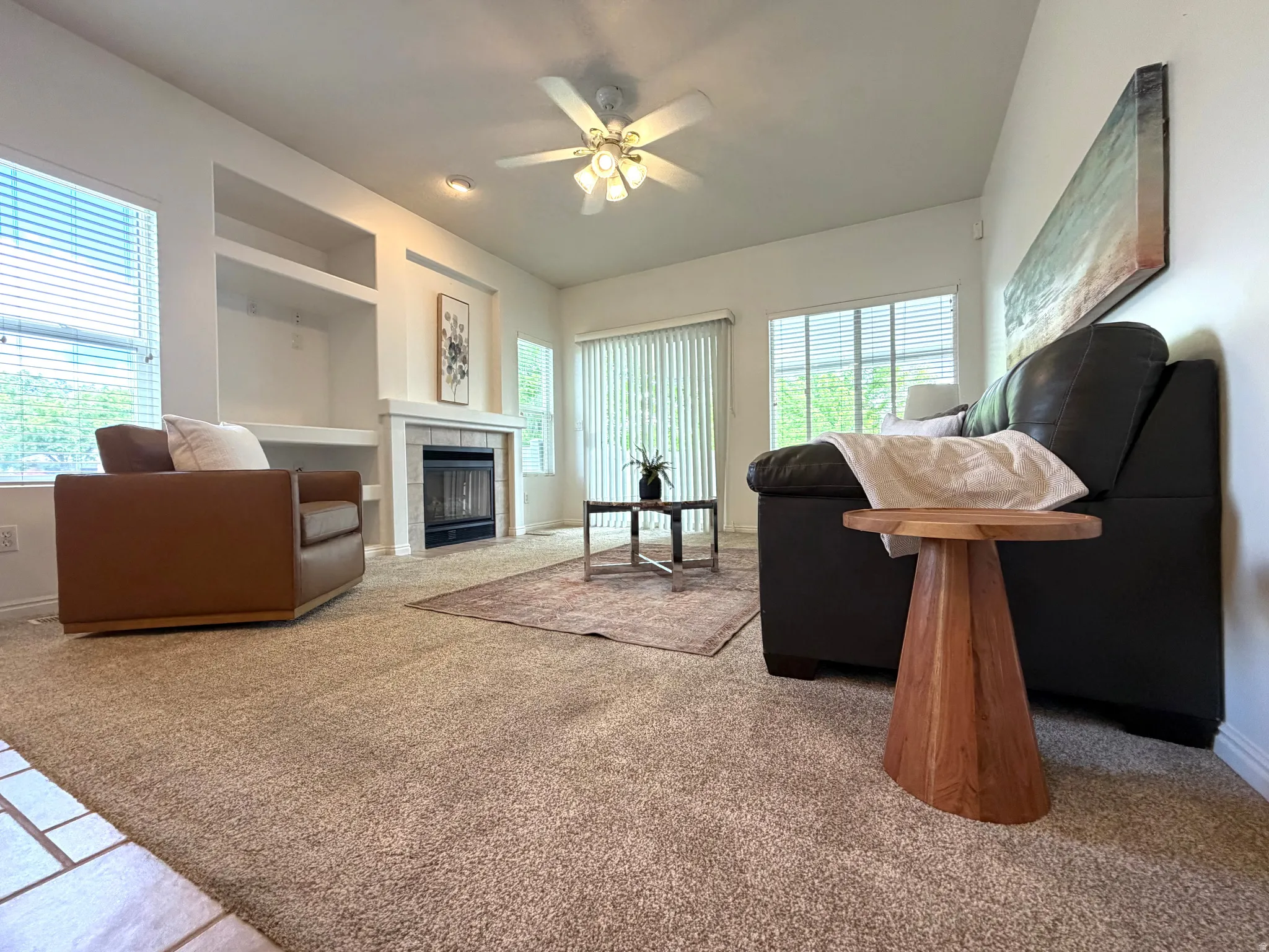 Living area featuring a central fireplace with light-toned tile surround and mantle, built-in shelving, windows with blinds, and ceiling fan with integrated lighting