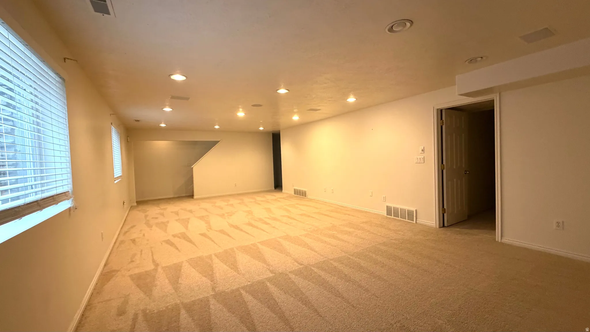 Spacious room with recessed lighting, patterned carpeting, multiple windows with blinds, a staircase, and a white paneled door