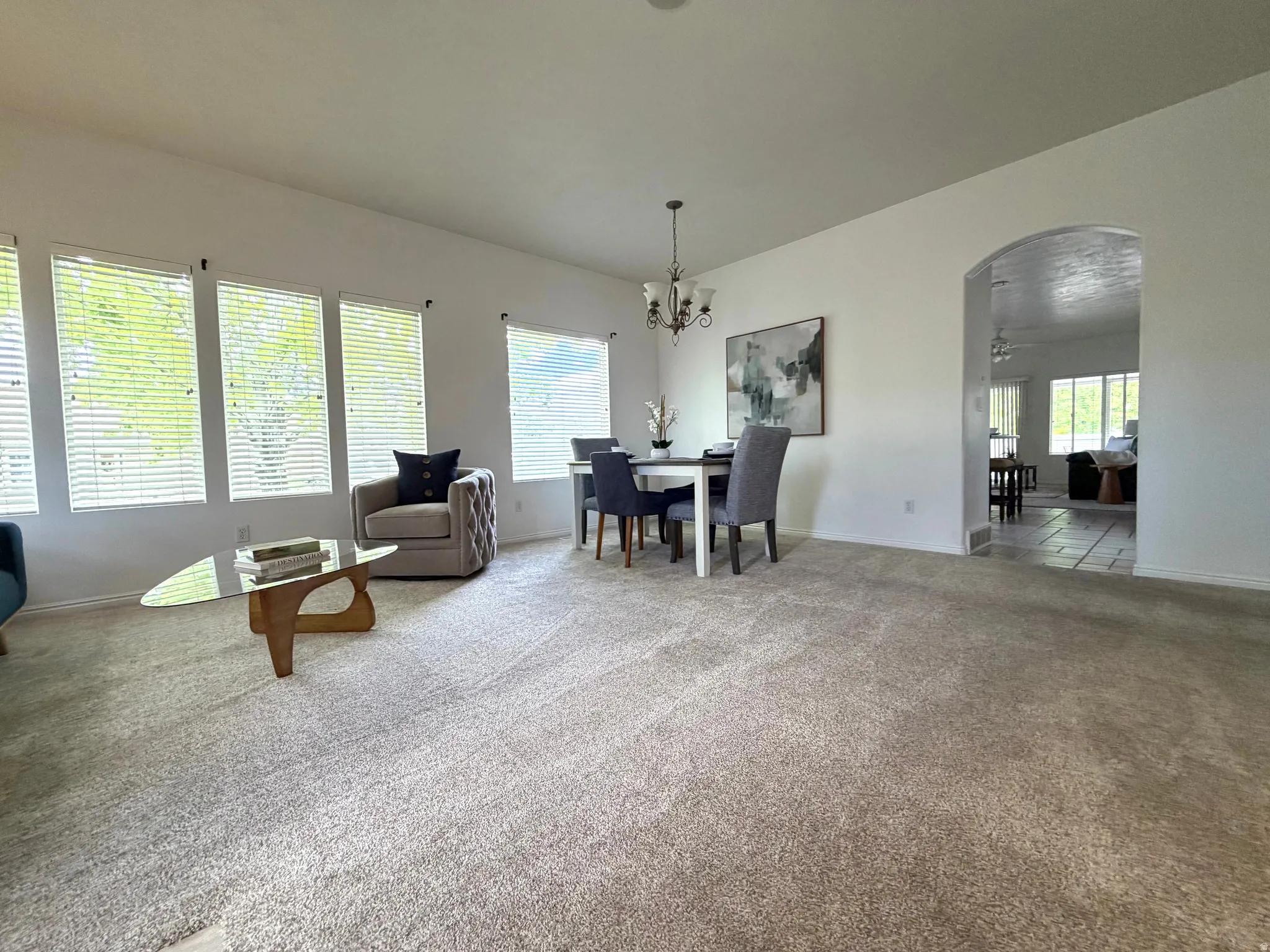 Spacious carpeted room featuring multiple windows with blinds, a dining area with a chandelier, and an arched doorway opening to a tiled area