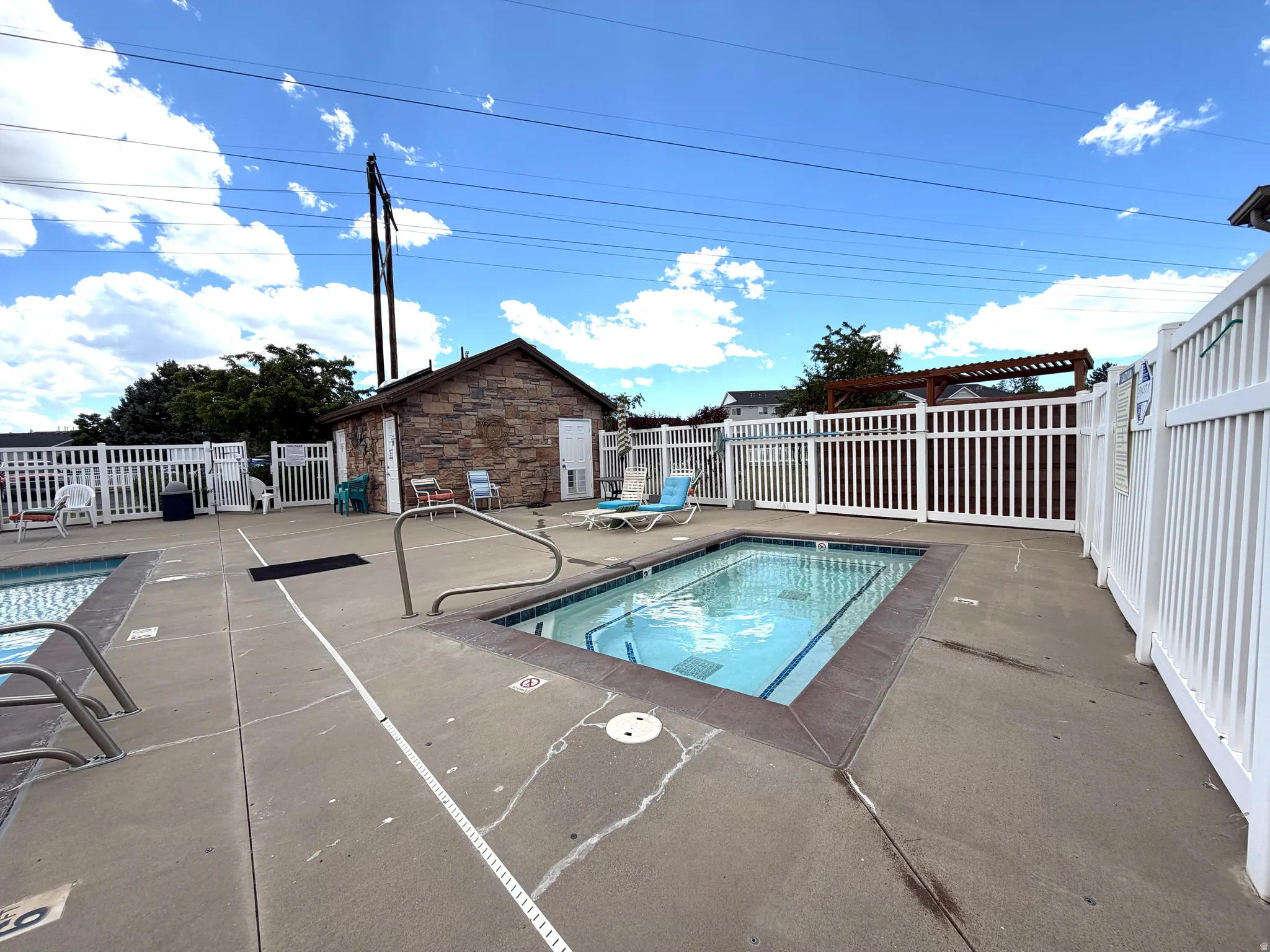 Concrete pool deck featuring two rectangular pools, white picket fencing, and a stone-clad utility building