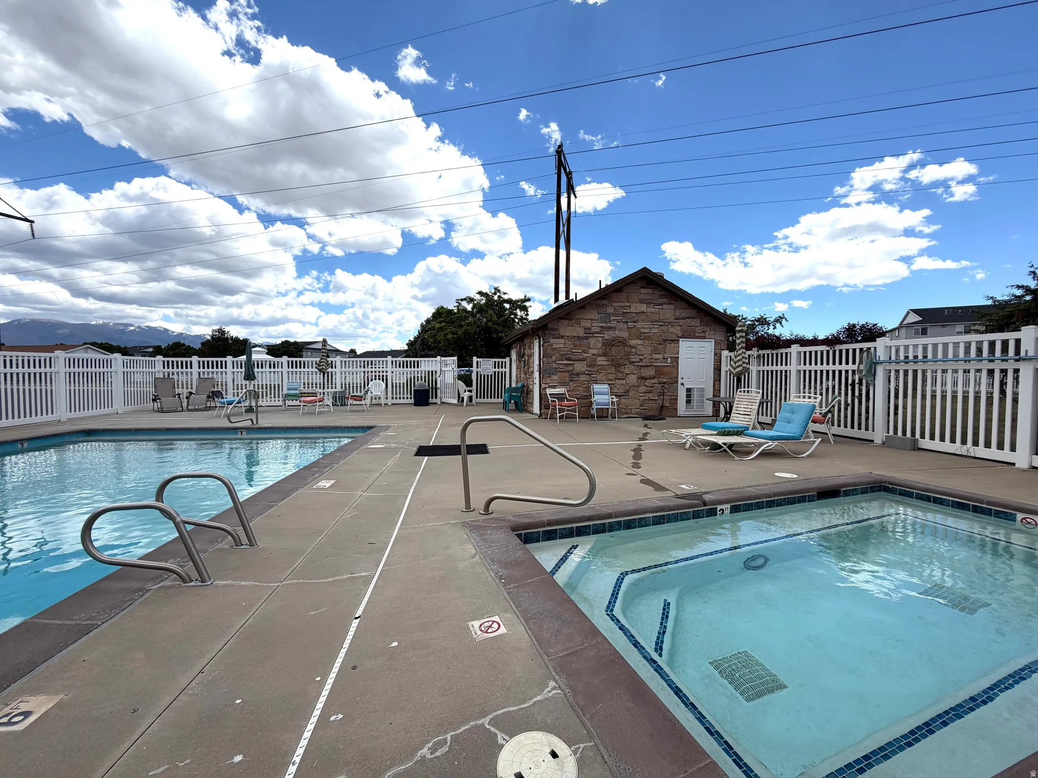 Outdoor swimming pool and spa area featuring a stone-clad building, white picket fencing, and concrete decking