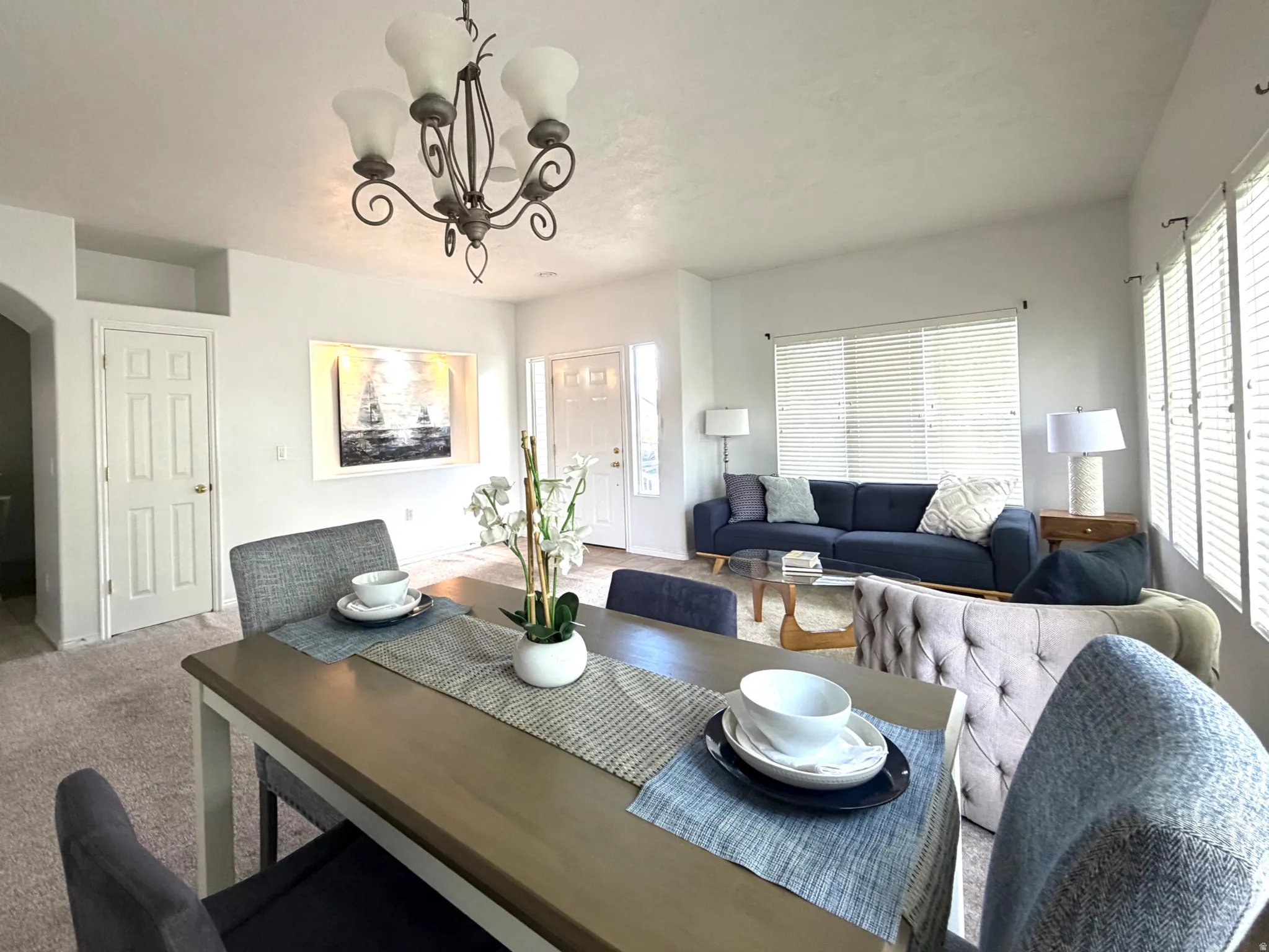 Open-concept living area with a decorative chandelier, recessed wall niche, and multiple windows with horizontal blinds
