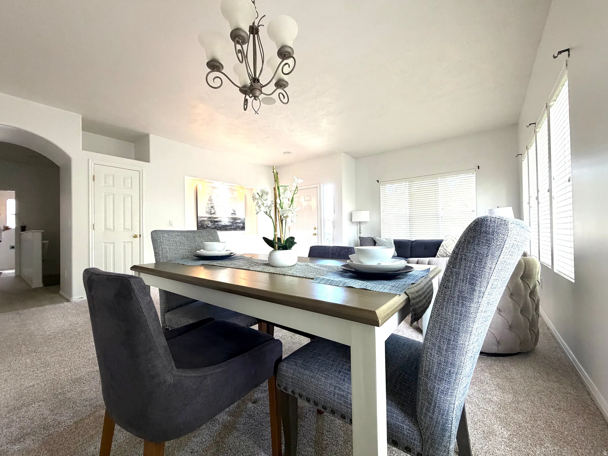 Open-concept living area featuring a dining space with a decorative chandelier, neutral carpet flooring, and multiple windows with blinds