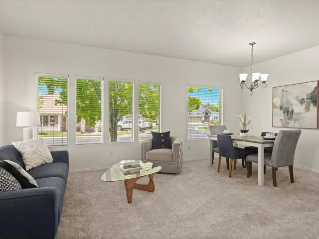 Bright living area featuring multiple large windows with blinds, neutral carpet flooring, and a chandelier over the dining space