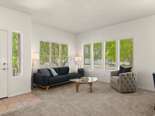 Living area featuring multiple windows with blinds, light-toned carpeting, and white painted walls