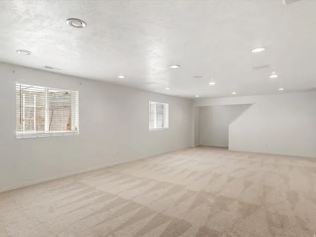 Spacious room featuring neutral carpeting, white walls, multiple windows with blinds, recessed lighting, and a visible staircase